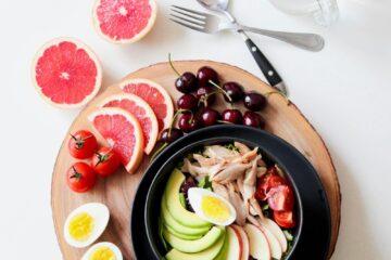 A vibrant breakfast flat lay featuring a mix of fruits, avocado, eggs, tomatoes, and sliced meats on a wooden platter.